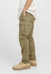 Olivegrüne Baumwoll-Cargo-Hosen mit Seitentaschen und schmalem Schnitt. Getragen mit beigen Schuhen mit dicker Sohle und Schnürsenkeln.