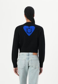 Black cropped sweater with ribbed hem, featuring a large blue heart pattern and text on the back. Worn with light blue jeans.