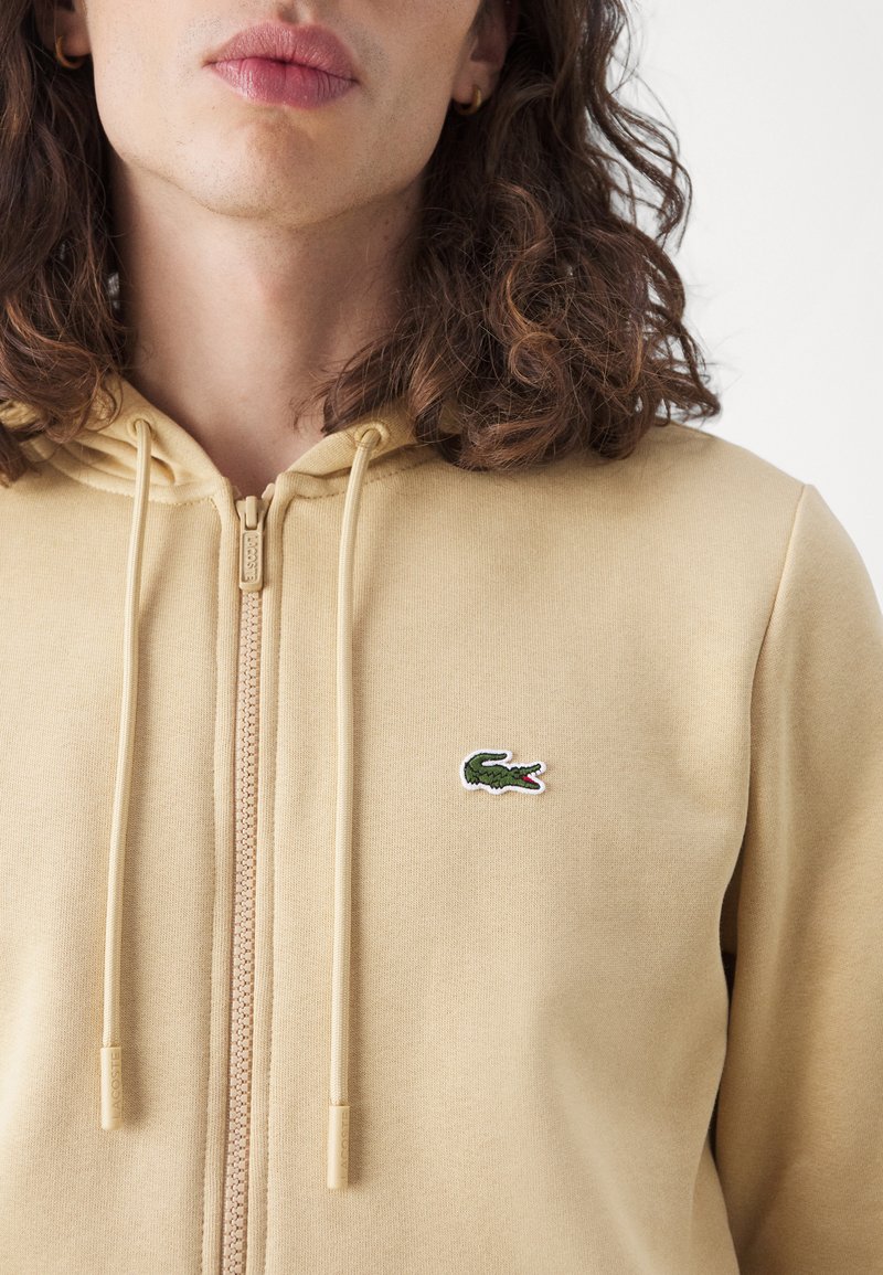 Beige zip-up hoodie met een kangoeroezak, trekkoordcapuchon en geborduurd krokodillenlogo op de borst. Zachte textuur en aansluitend ontwerp.