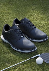 Chaussures de golf noires avec semelles blanches sur l’herbe à côté d’un club de golf et d’une balle de golf blanche.