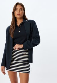 Blazer de marina con un solo botón, superpuesto a una camiseta polo negra con botones dorados, combinado con una falda mini a rayas en azul marino y crema.
