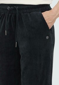 Zwarte corduroy broek met verticale ribbelstructuur, elastische taille met trekkoord, zijzakken en een klein zilveren logo accent.