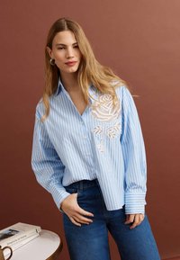 Kobieta o długich blond włosach, ubrana w niebieską koszulę w paski z białą koronką w kwiaty oraz niebieskie dżinsy, stojąca obok stołu z książkami.