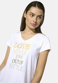 Weißes V-Ausschnitt-T-Shirt aus weichem Material mit Text in verschiedenen Farben: "LIEBE WIRD NIE OUT OF STYLE SEIN." Kurze Ärmel und lockere Passform.