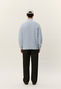 Mężczyzna ubrany w jasnoniebieski dzianinowy sweter, czarne spodnie, czarne buty oraz białą czapkę z daszkiem, stoi tyłem na jednolitym tle.