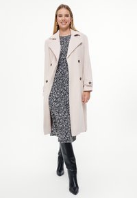 Jasnoróżowy płaszcz o podwójnym zapięciu, z brązowymi guzikami i dużymi klapami, noszony na czarno-białej wzorzystej sukience oraz czarnych kozakach do kolan.