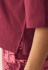 Burgundi värvi T-särk, millel on külgauk, lühikesed varrukad ja pehme puuvilla tekstuur. Allpool on mustriline kangas roosa ja tumepunase toonides.