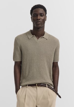 Man wearing a textured beige short-sleeve polo shirt tucked into beige pants with a braided brown belt, hands in pockets, neutral background.