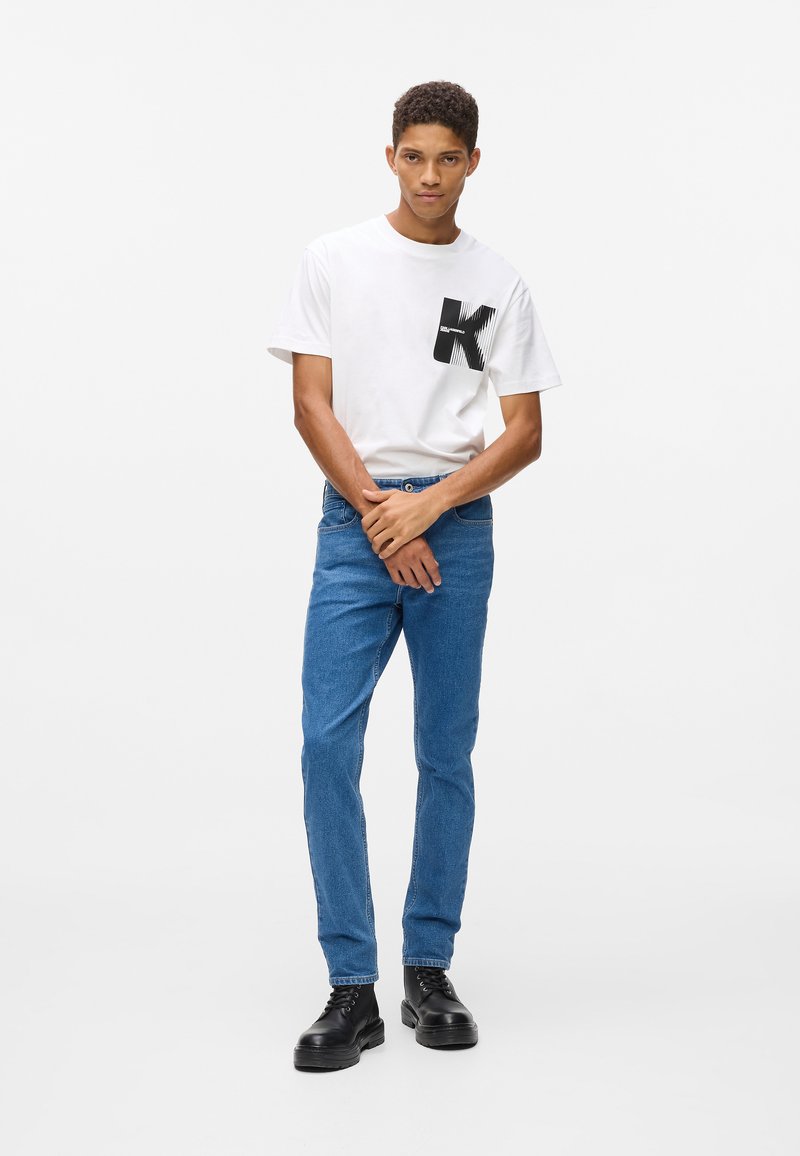 Jauns vīrietis stāv balta fona priekšā, tērpies baltā T-kreklā ar melnu K logos, zilu džinsu biksēs un melnās šņorējamās kurpēs.