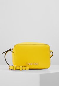 Borsa a tracolla in pelle gialla di forma rettangolare, con il marchio "Calvin Klein" in oro, dotata di tracolla removibile e superficie testurizzata.