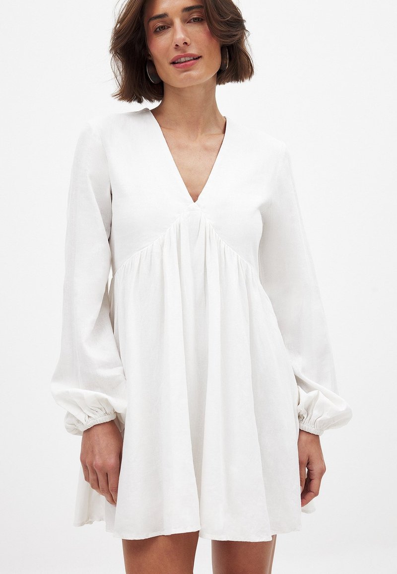 Weißes Leinenkleid mit V-Ausschnitt, lockerer Passform und gerafften Details. Lange Ärmel mit elastischen Bündchen. Glatte Textur, knielang.