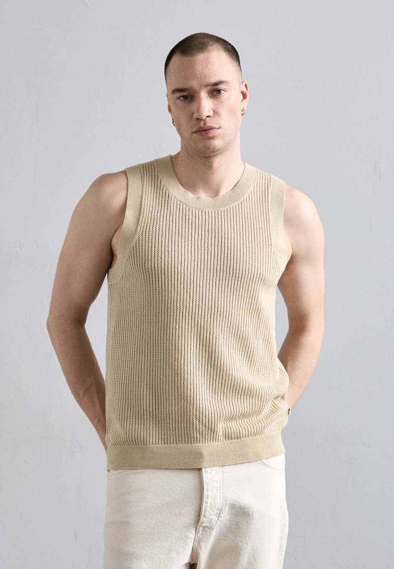 Joven con cabello corto, vistiendo un chaleco de punto sin mangas beige y pantalones claros, de pie frente a una pared lisa de color gris claro.