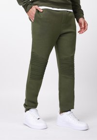 Pantaloni sport verzi măsliniu cu accente nervurate la genunchi, talie elastică și buzunare laterale, asortați cu pantofi sport albi.