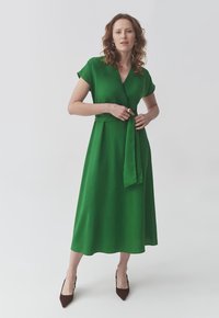 Rochie verde înfoiată, cu mâneci scurte, decolteu în V și curea în talie. Țesătură netedă și fluidă, până la genunchi, asociată cu pantofi cu vârful ascuțit, maro.