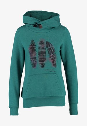Persona che cammina indossando una felpa con cappuccio verde acqua con motivo a penna, jeans chiari sbiaditi e sneakers Adidas nere con motivo.