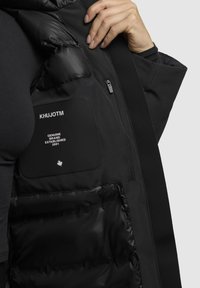 Doudoune noire avec une doublure intérieure brillante et matelassée. Comprend une poche zippée et une étiquette avec "KHUJOTM" et "Marque Authentique Établie en 2001."