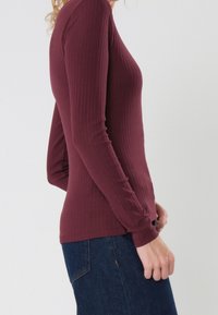 Burgundy long-sleeve top with ribbed texture. Form-fitting design, rounded neckline, and fitted cuffs. Paired with dark blue jeans.