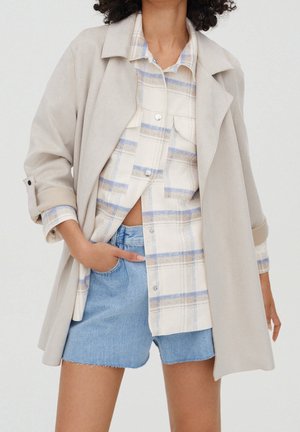 Frau trägt einen hellbeigen Mantel über einem beigen und blauen karierten Hemd und hellblauen Jeansshorts, Hand in der Tasche, vor einfachem Hintergrund.