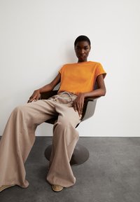 T-shirt orange à manches courtes avec un petit logo, associé à un large pantalon beige, assis sur une chaise sombre et courbée, contre un fond neutre.