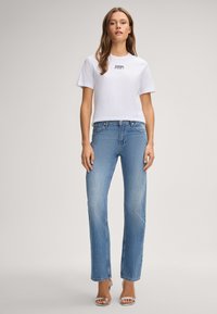 Weiße Baumwoll-T-Shirt mit "JOOP!"-Text, kombiniert mit hellblauen geraden Jeans und metallic-silbernen offenen Absätzen. Glatte Texturen überall.