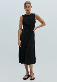 Schwarzes ärmelloses Kleid mit einem geknoteten Detail an der Taille, knielang, glatte Textur und entspannter Schnitt, kombiniert mit schwarzen Sandalen.