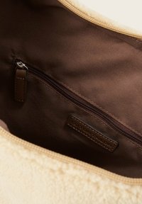 Wnętrze torebki w kolorze kremowym i brązowym z zamykaną na zamek kieszonką z skórzaną zawieszką oraz materiałową metką z logo. Miękka, teksturowana zewnętrzna powierzchnia.