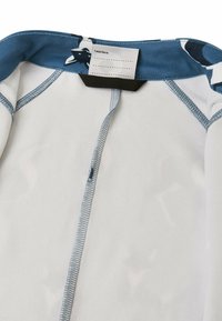 Weißes Stoffhemd mit blauem Rundhalsausschnitt, sichtbaren Nähten und einem Etikett. Enthält ein gemustertes Kragen-Design mit Sternen und Akzenten.