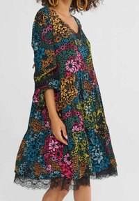Femme portant une robe patchwork fleurie multicolore avec manches retroussées et volant en dentelle à l'ourlet.