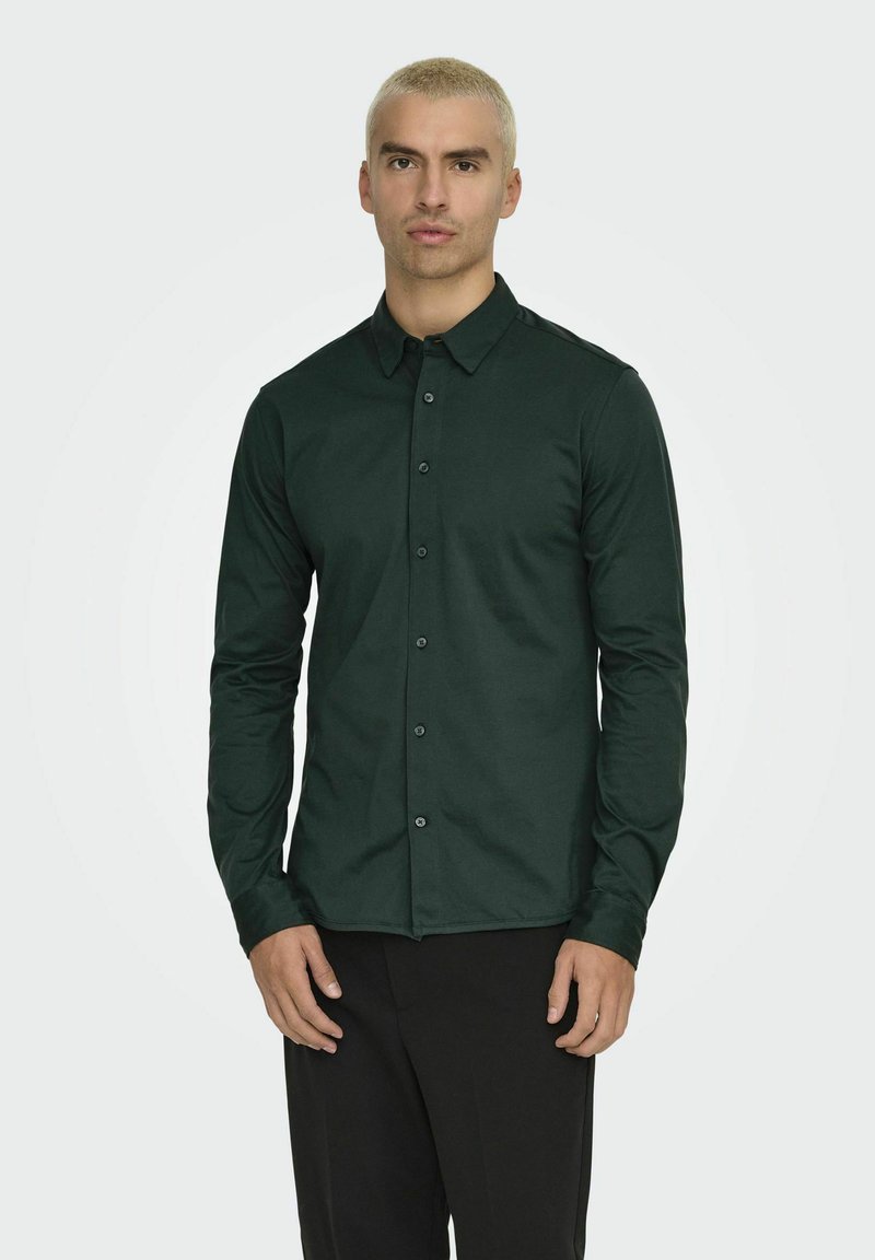 Camisa de botón verde oscuro con mangas largas, cuello y una textura suave. La camisa tiene un diseño ajustado y botones negros.