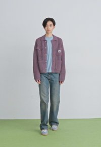 Junge Person steht auf grünem Boden, trägt einen lila gestreiften Cardigan, ein hellblaues Shirt, blaue Jeans und graue Sneaker vor einem einfarbigen Hintergrund.