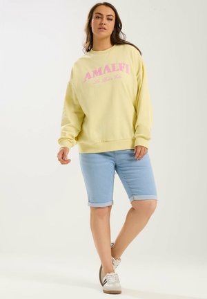 Frau trägt ein gelbes Sweatshirt mit der Aufschrift "AMALFI La Dolce Vita", hellblaue Jeansshorts und weiße Sneaker, steht vor einem schlichten Hintergrund.