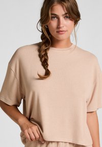 Licht beige cropped sweatshirt van zachte stof, met korte mouwen en een ronde halslijn, lichtjes vastgehouden door een hand.