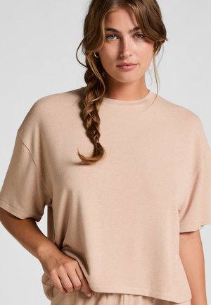 Helles beiges, kurz geschnittenes Sweatshirt aus weichem Stoff, mit kurzen Ärmeln und rundem Ausschnitt, leicht von einer Hand gehalten.