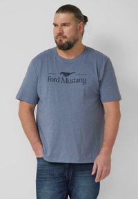 Niebieska bawełniana koszulka z czarną grafiką Forda Mustanga i napisem. Koszulka ma luźny krój i krótkie rękawy, zestawiona z niebieskimi dżinsami.