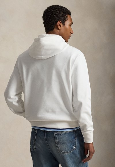 Άνδρας με την πλάτη γυρισμένη, φοράει λευκό φούτερ με κουκούλα και μπλε τζίν μέσα σε ουδέτερο μπεζ φόντο.