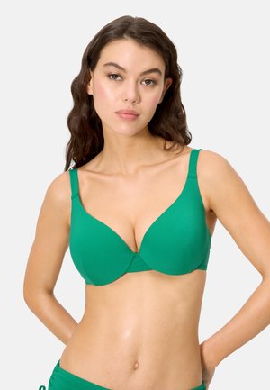 Joven con cabello largo y ondulado, vistiendo un sujetador con aro verde y braguitas a juego, de pie contra un fondo claro y liso.