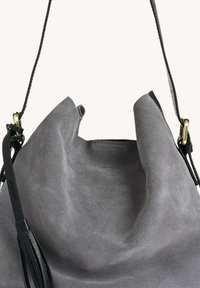Sac seau en suede gris avec un haut replié, une sangle en cuir noir et des finitions dorées. Caractéristiques d'une texture lisse et douce avec un minimalisme dans les détails.