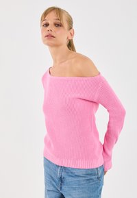 Donna con capelli biondi che indossa un maglione rosa senza spalline e jeans blu, in piedi su uno sfondo bianco uniforme.