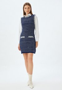 Tummansininen tweed-mekko, jossa on ruudullinen kuvio, risaset reunat ja kaksi etutaskua. Päällä valkoinen kauluspaita.