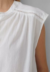 Chemise en lin blanc à manches courtes, avec un système de fermeture à boutons et de délicates dentelles le long de la couture de l'épaule. Tissu doux et texturé.