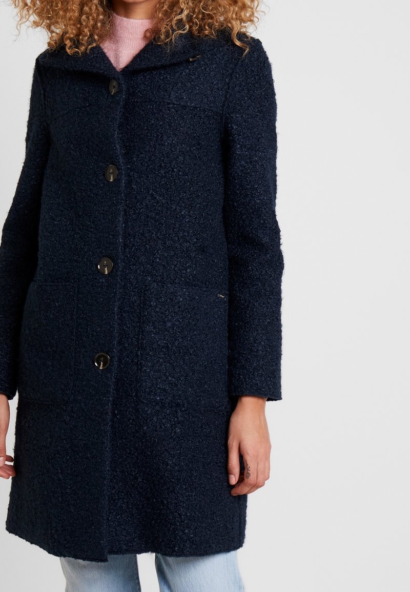 Cappotto in lana strutturata blu navy con grandi bottoni frontali, colletto che valorizza, tasche laterali e vestibilità dritta. Presenta una leggera lucentezza.