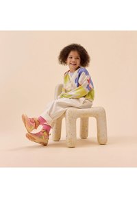 Child sits on a speckled white chair, wearing a multi-colored sweater, cream pants, and pink socks, with pink and yellow sneakers.