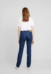 Vaqueros de mezclilla azul con un corte recto, con una etiqueta de marca amarilla en la parte trasera, combinados con una camiseta blanca y zapatos blancos.