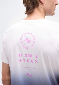Άτομο που φοράει λευκό μπλουζάκι με μπλε διαβάθμιση και ροζ κείμενο "MY SPORT IS HYROX" και λογότυπο αγώνων στην πλάτη.