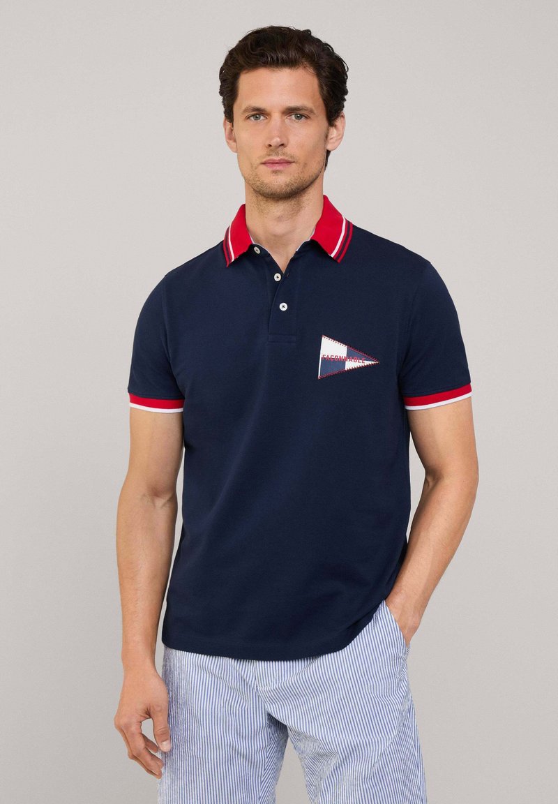 Granatowa koszulka polo z czerwonym kołnierzykiem i akcentami na rękawach. Posiada geometryczny wzór łatki z białymi i czerwonymi detalami. Wytrzymały materiał.