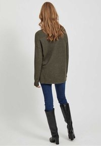 Femme aux longs cheveux ondulés portant un pull vert olive, un jean skinny bleu et des bottes noires montantes, debout de dos sur un fond blanc.