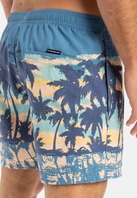 Badeshorts in Blau mit einem Palmenmuster in Gelb und Marineblau. Ausgestattet mit einer hinteren Gesäßtasche mit Druckknopfverschluss und Logopatch.