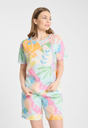 Vrouw draagt een kleurrijk pastel abstract print shirt met korte mouwen en bijpassende shorts, kijkt opzij tegen een witte achtergrond.