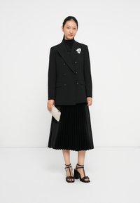 Blazer nero doppiopetto sopra una gonna plissettata nera. Gli accessori includono una spilla a forma di fiore in argento e una pochette bianca testurizzata. Tacchi neri con cinturini.