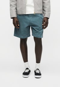 Άνδρας που φοράει πετρόλ βερμούδα από κορντόρι, λευκές κάλτσες, μαύρα αθλητικά παπούτσια, λευκό πουκάμισο και ανοιχτό γκρι σακάκι, στέκεται μπροστά σε απλό φόντο.